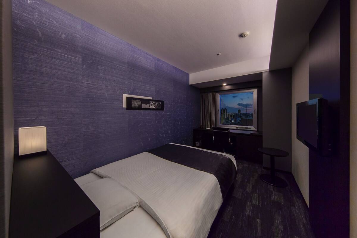 A hotel room with a large bed, a desk, a chair, a TV, and a window with a city view.