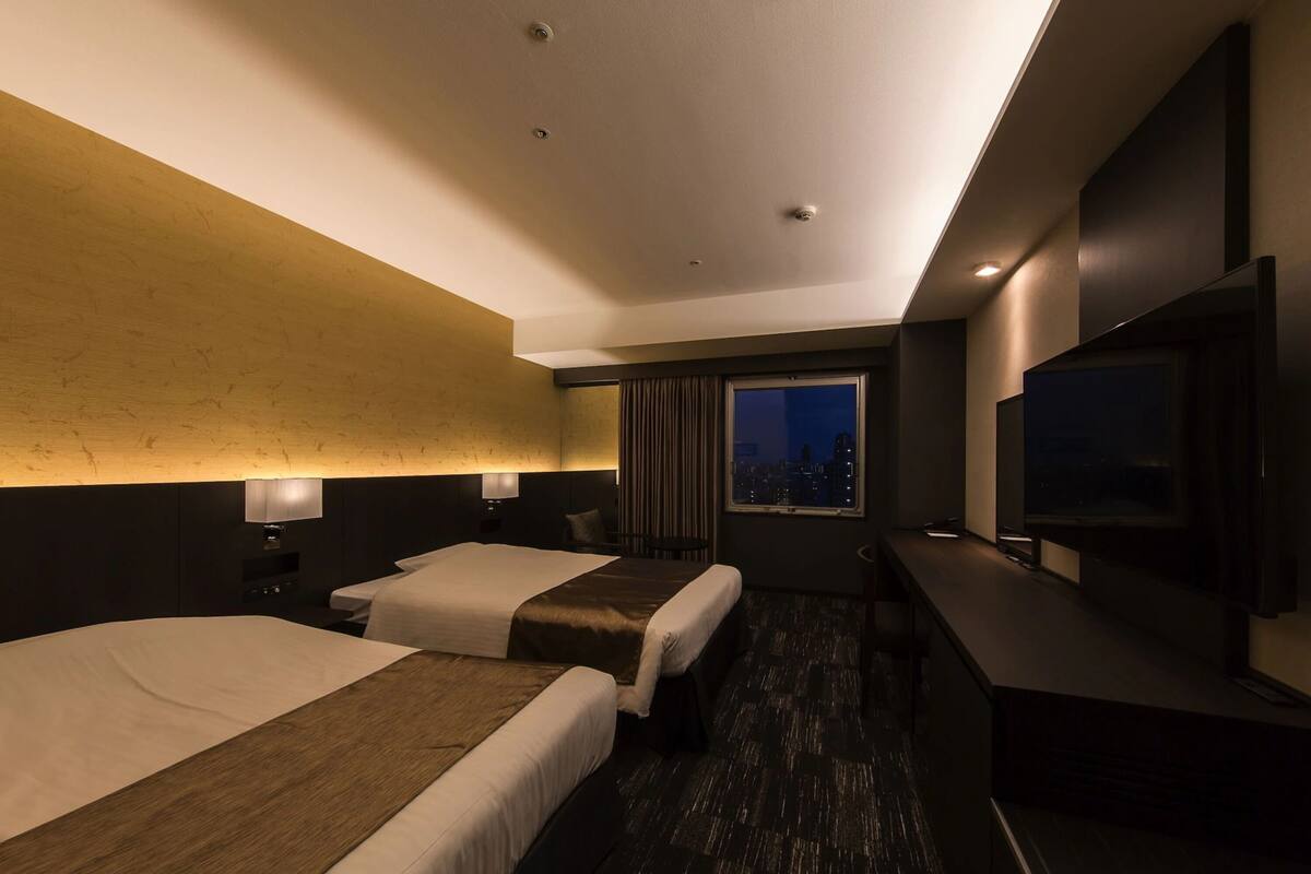A hotel room with two beds, a TV, a desk, and a city view.