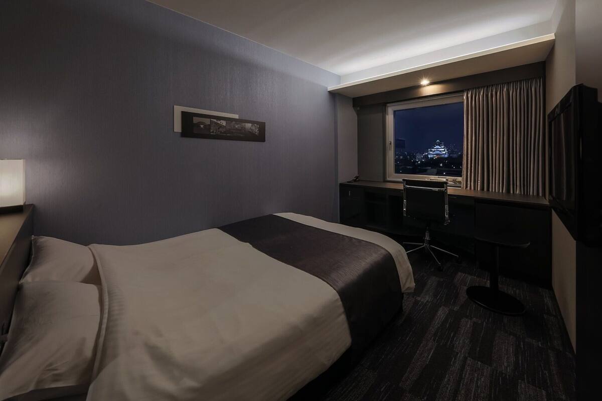 A hotel room with a large bed, a desk, a chair, a TV, and a window with a city view.
