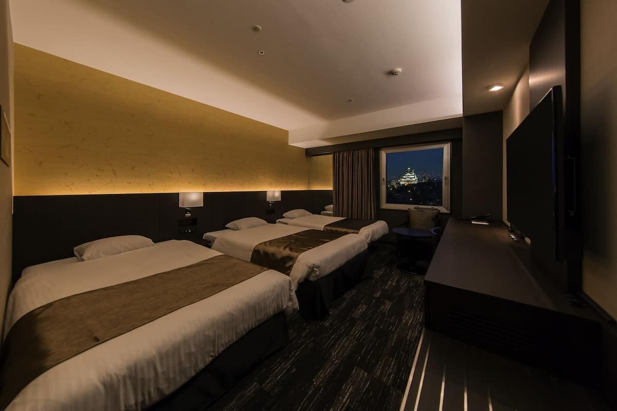 A hotel room with three beds, a large window with a city view, a flat-screen TV, and a projector.