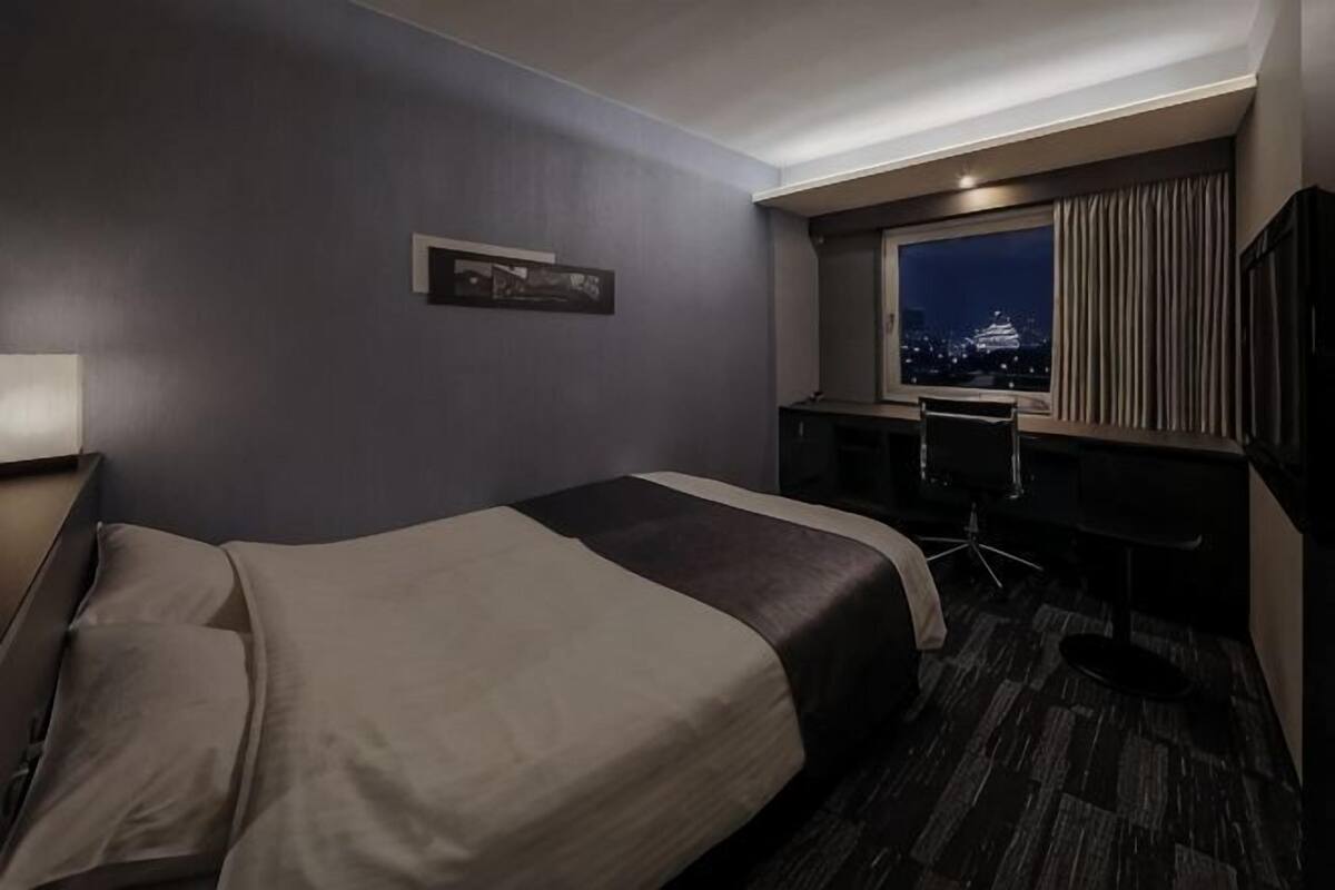 A hotel room with a bed, desk, chair, and a view of a cityscape at night.