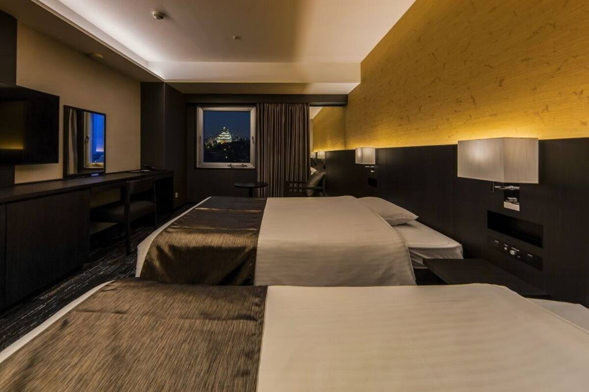 A hotel room with two beds, a TV, a desk, and a window with a view of a castle.