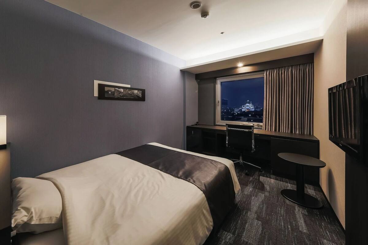 A hotel room with a bed, a desk, a chair, a lamp, and a window with a cityscape view.