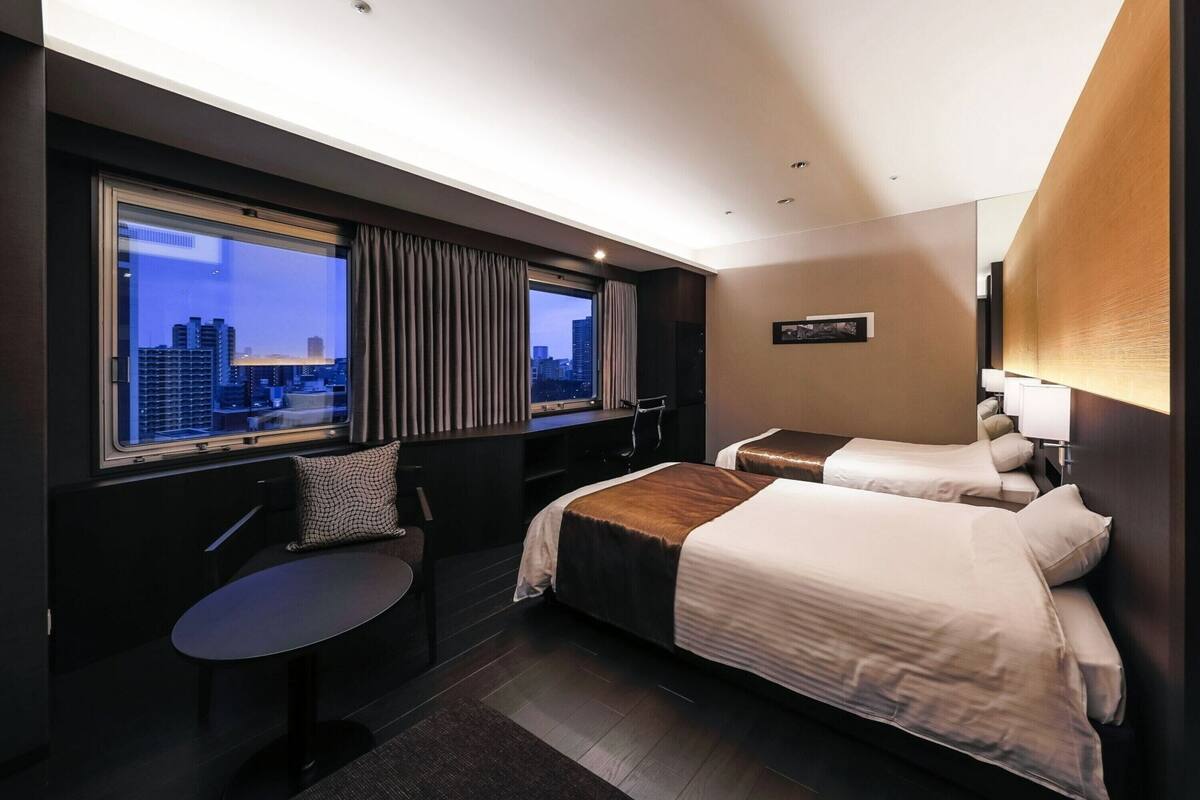 A hotel room with two beds, a desk, and a chair. There is a large window with a city view.