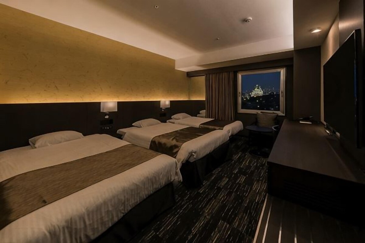 A hotel room with three beds, a large window with a city view, a desk, and a flat-screen TV.