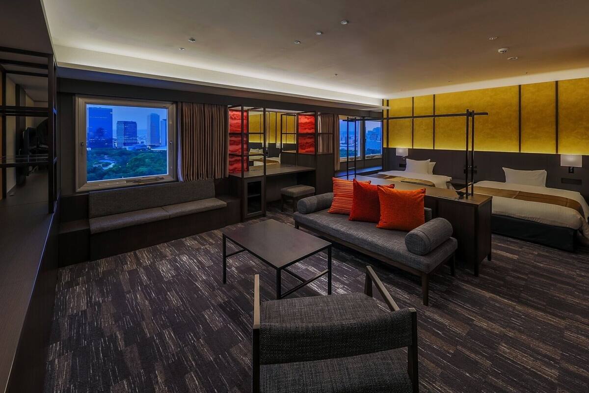 A modern hotel room with a sofa, two beds, a desk, and a view of the cityscape.