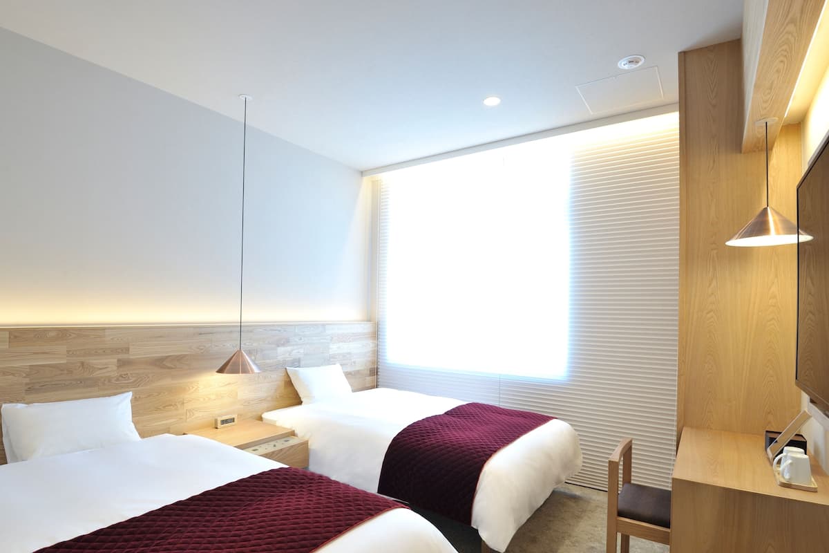 Habitación de hotel con dos camas, cabecero de madera, mesita de noche con lámpara, ventana con persianas y escritorio de madera con una silla.