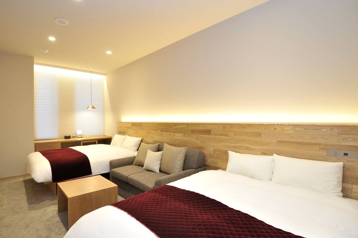 Una habitación de hotel moderna con dos camas, un sofá, una mesita de madera y una ventana con persianas.