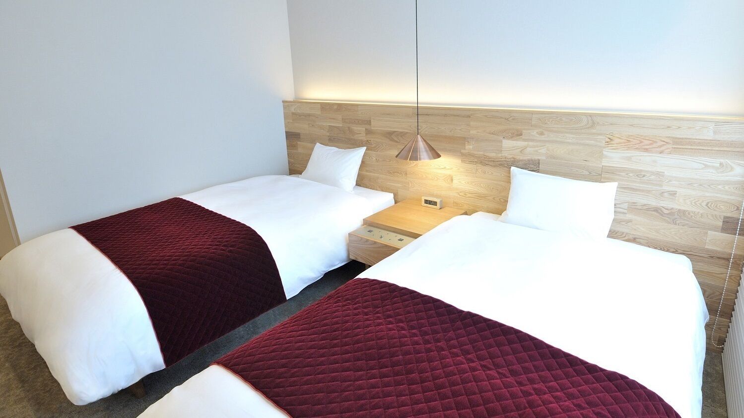 Twee bedden met wit en rood beddengoed, een houten hoofdeinde, een nachtkastje met een lamp en een wandklok.