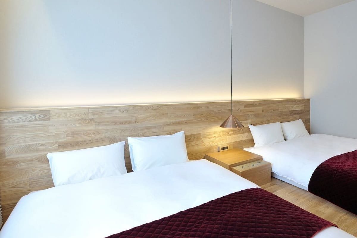 Habitación de hotel con dos camas, cabecero de madera, mesita de noche con lámpara y luz empotrada en la pared.