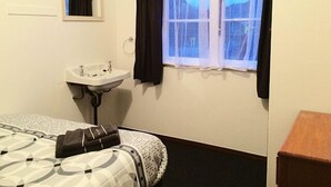 Een kleine, schone kamer met een bed, een raam met gordijnen, een wastafel en een spiegel.