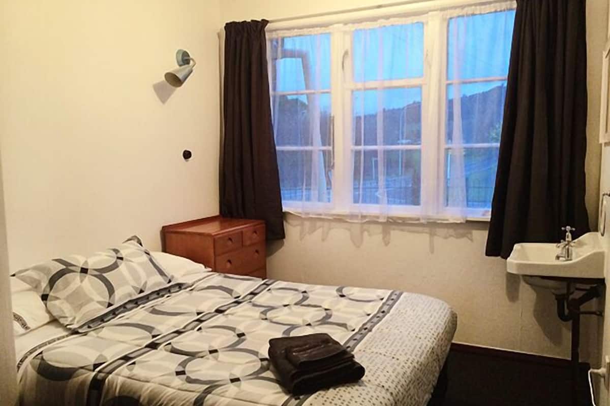 A bedroom with a bed, a nightstand, a window with curtains, and a bathroom with a sink.