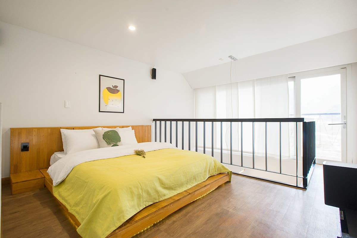 A bedroom with a wooden bed, a yellow bedspread, a white pillow, and a green pillow. There is a black railing and a window with a view.
