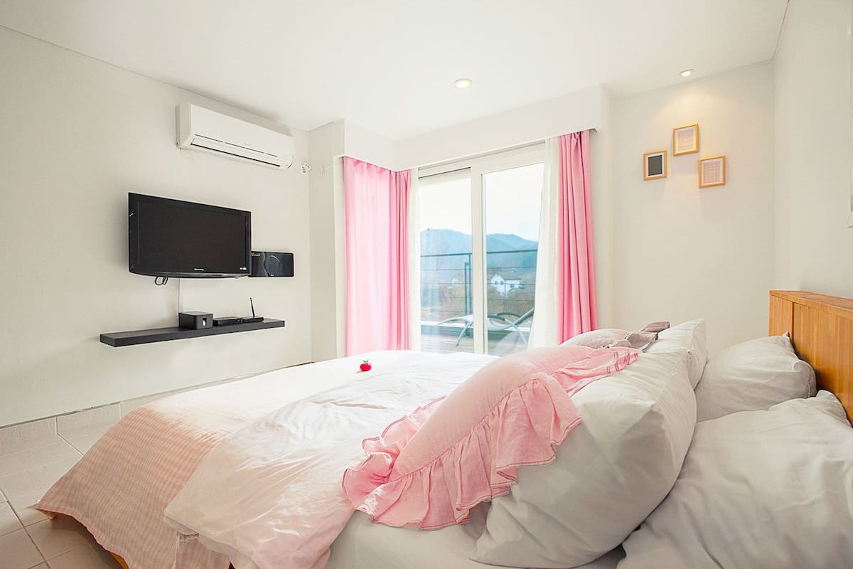 A bedroom with a bed, a TV mounted on the wall, a window with curtains, and a view of the outdoors.