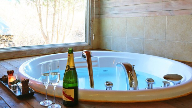 A hot tub with champagne and glasses, a window with a view of trees, and wooden walls. - Skylake Pension (Hongcheon)
