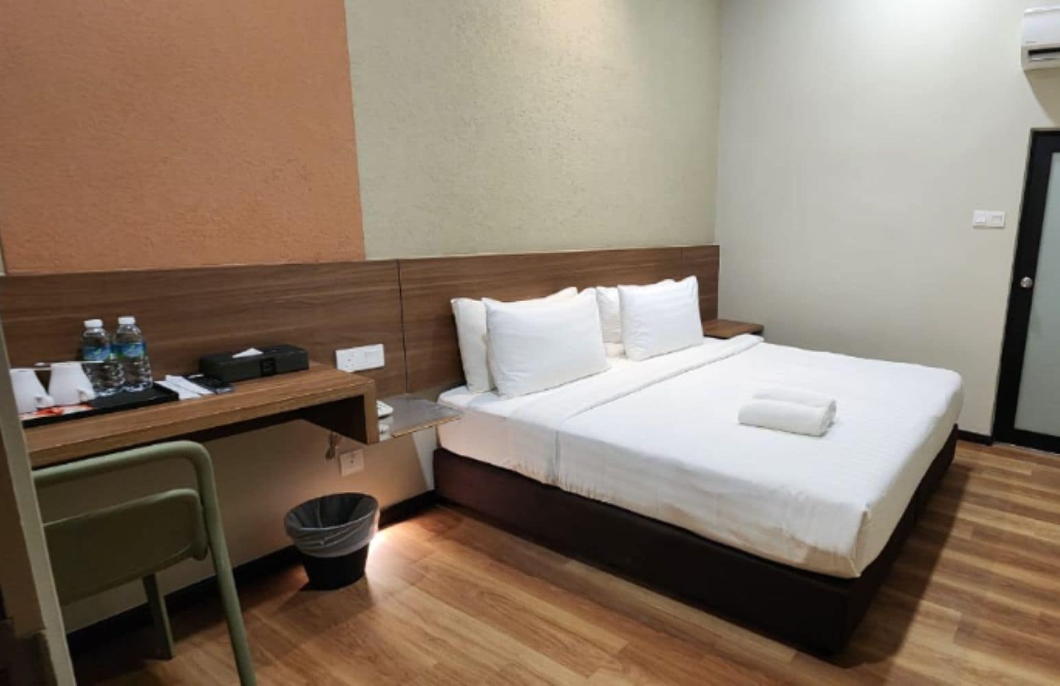 Habitación de hotel con cama, escritorio, silla, cubo de basura y unidad de aire acondicionado instalada en la pared.