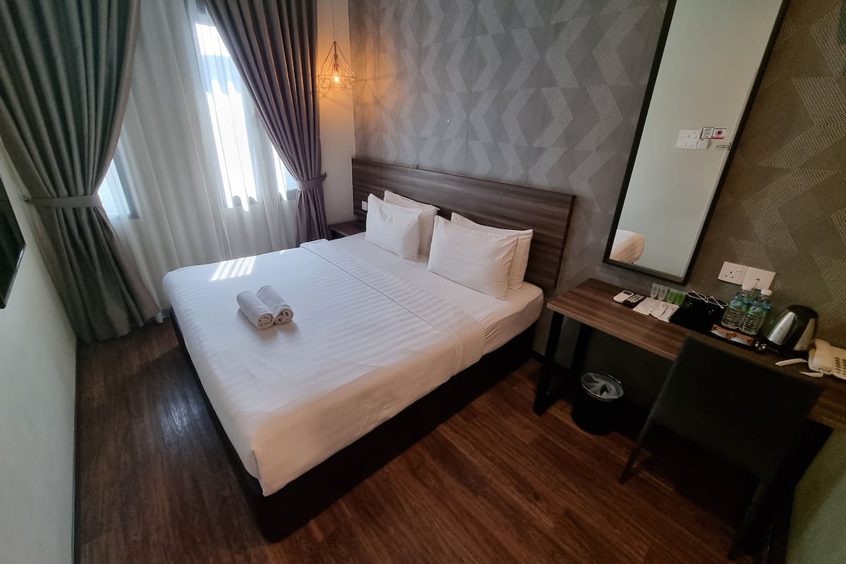 A hotel room with a neatly made bed, a bedside table with a lamp, a mirror, and a desk with a chair.
