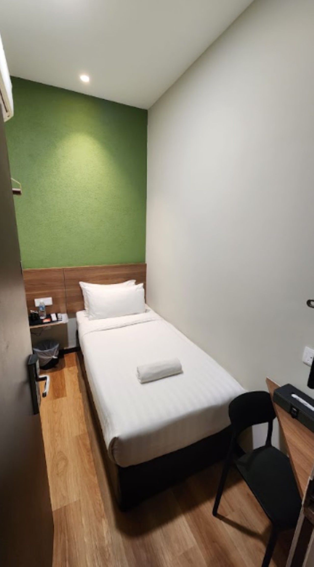 A small, single-bed room with a wooden headboard, a bedside table, a chair, and a desk with a computer.