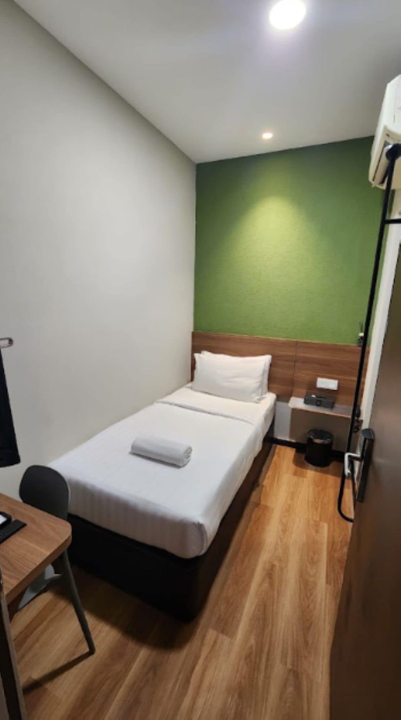 A small, neatly arranged bedroom with a single bed, a bedside table, a chair, and a green accent wall.