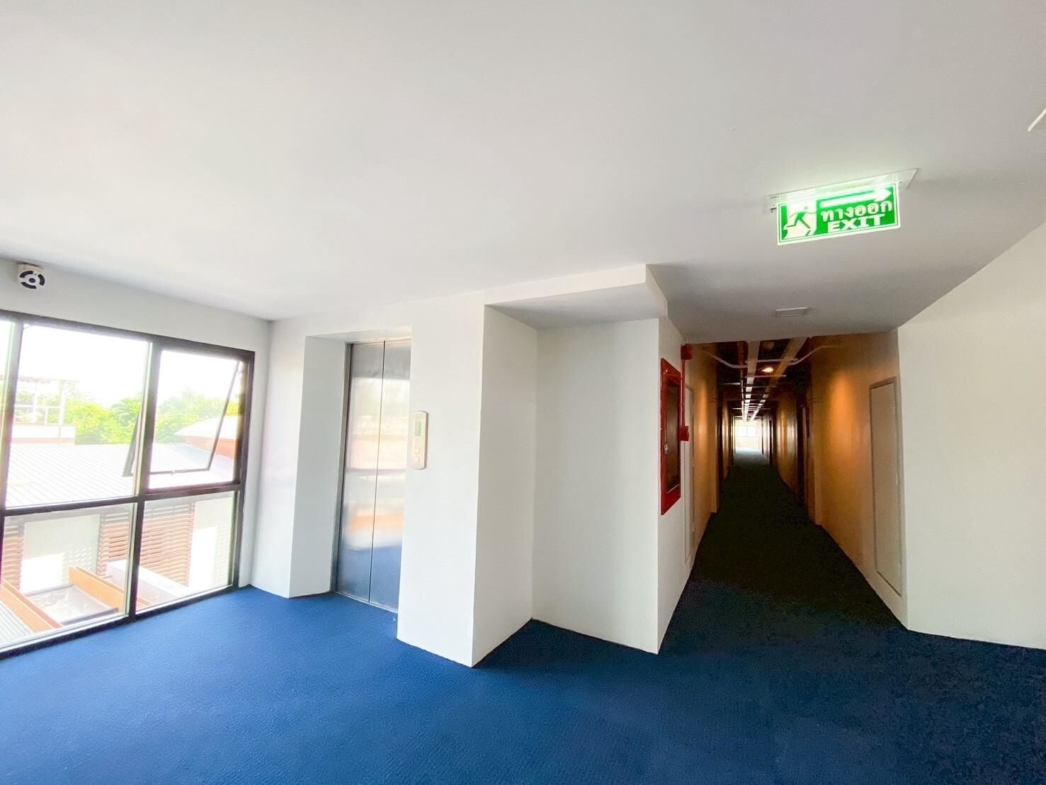 Un pasillo con alfombra azul, una señal de salida visible y una ventana con persianas.