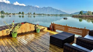 Una terraza de madera con mobiliario de mimbre, plantas en macetas y vistas a un lago tranquilo con montañas al fondo.