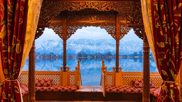 Una cama de madera tradicional con intrincados grabados, un tapete rojo estampado y vistas de montañas nevadas a través de una ventana ornamentada.