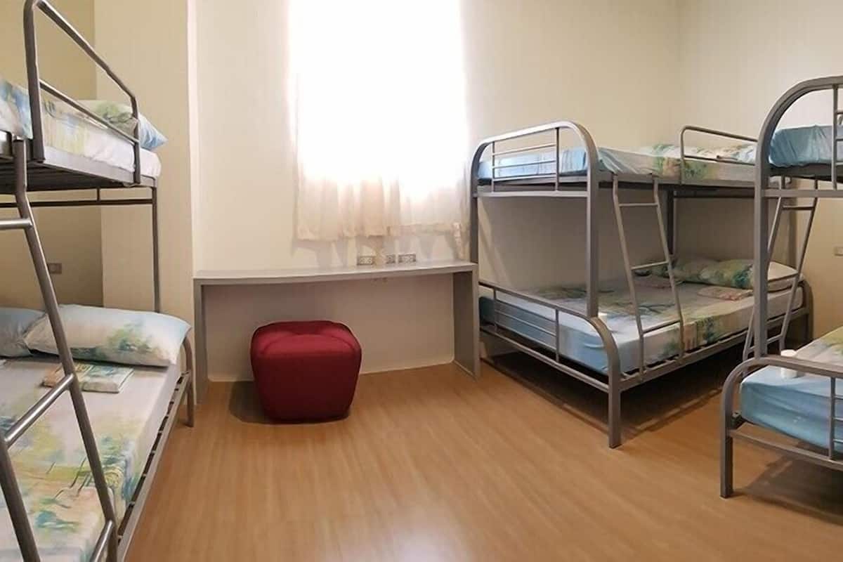 Quarto tipo dormitório com beliches, um pequeno banquinho vermelho e uma janela com cortinas.