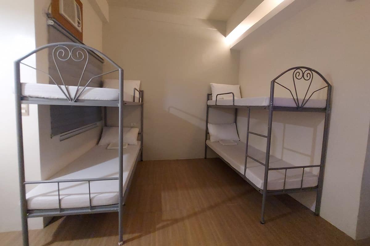 Quarto pequeno com duas camas beliches, piso de madeira e luminária no teto.