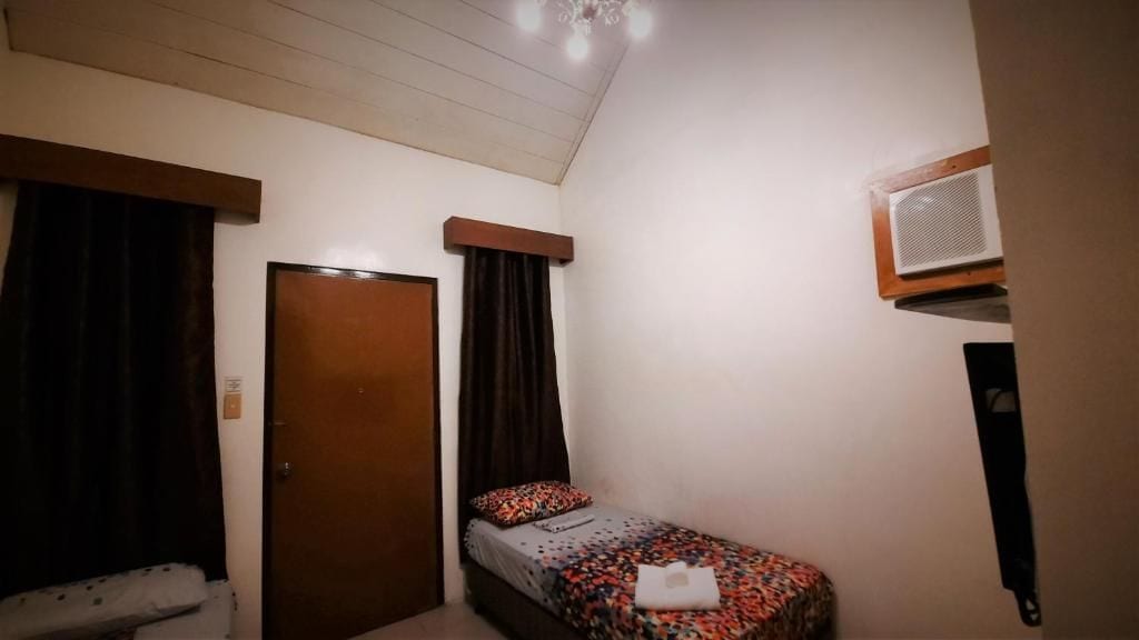 A small, single-bed room with a wooden door, a window with curtains, a bed with a floral bedspread, and a wall-mounted air conditioning unit.