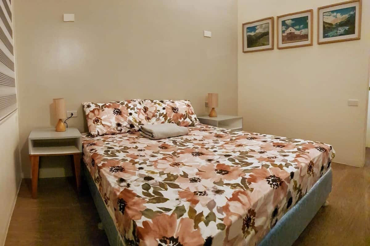 Quarto com cama, duas mesas de cabeceira, abajur, janela com persianas e três quadros na parede.