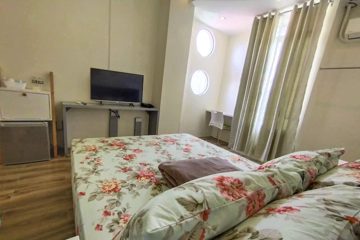 Quarto de hotel com cama, televisão, escrivaninha e janela com cortinas.