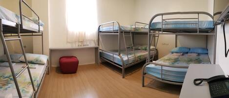 A dormitory room with bunk beds, a desk with a telephone, and a red ottoman.