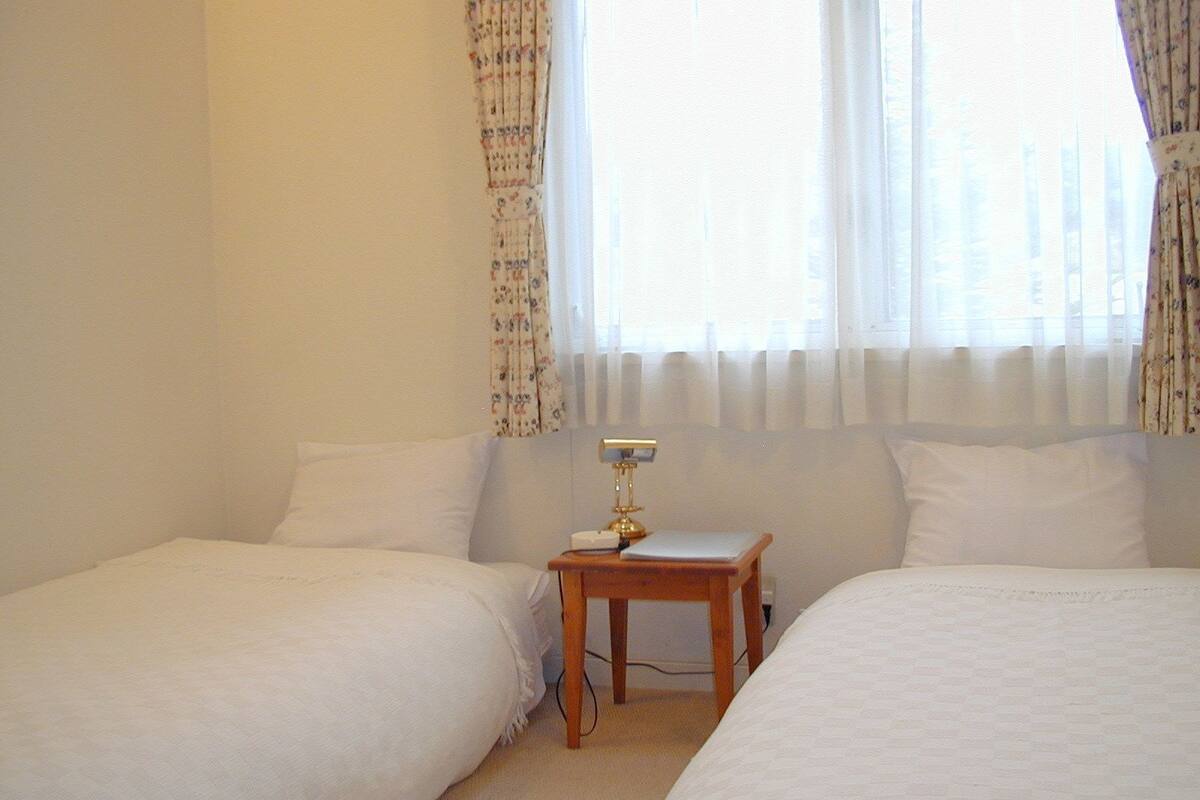 A hotel room with two single beds, a small wooden table with a lamp, and a window with sheer curtains.
