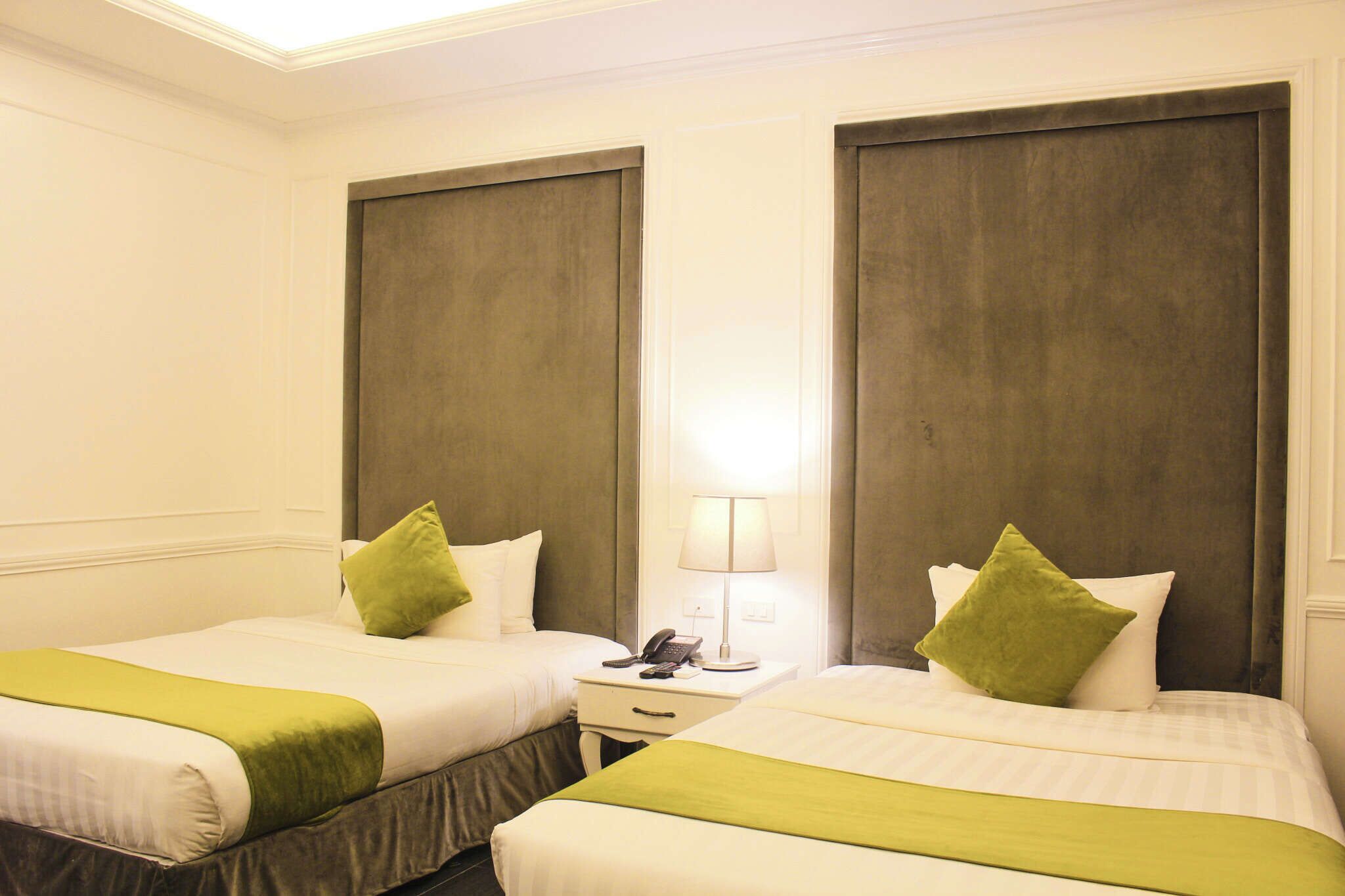 A hotel room with two beds, each with a green accent pillow, a bedside table with a lamp, and a phone.