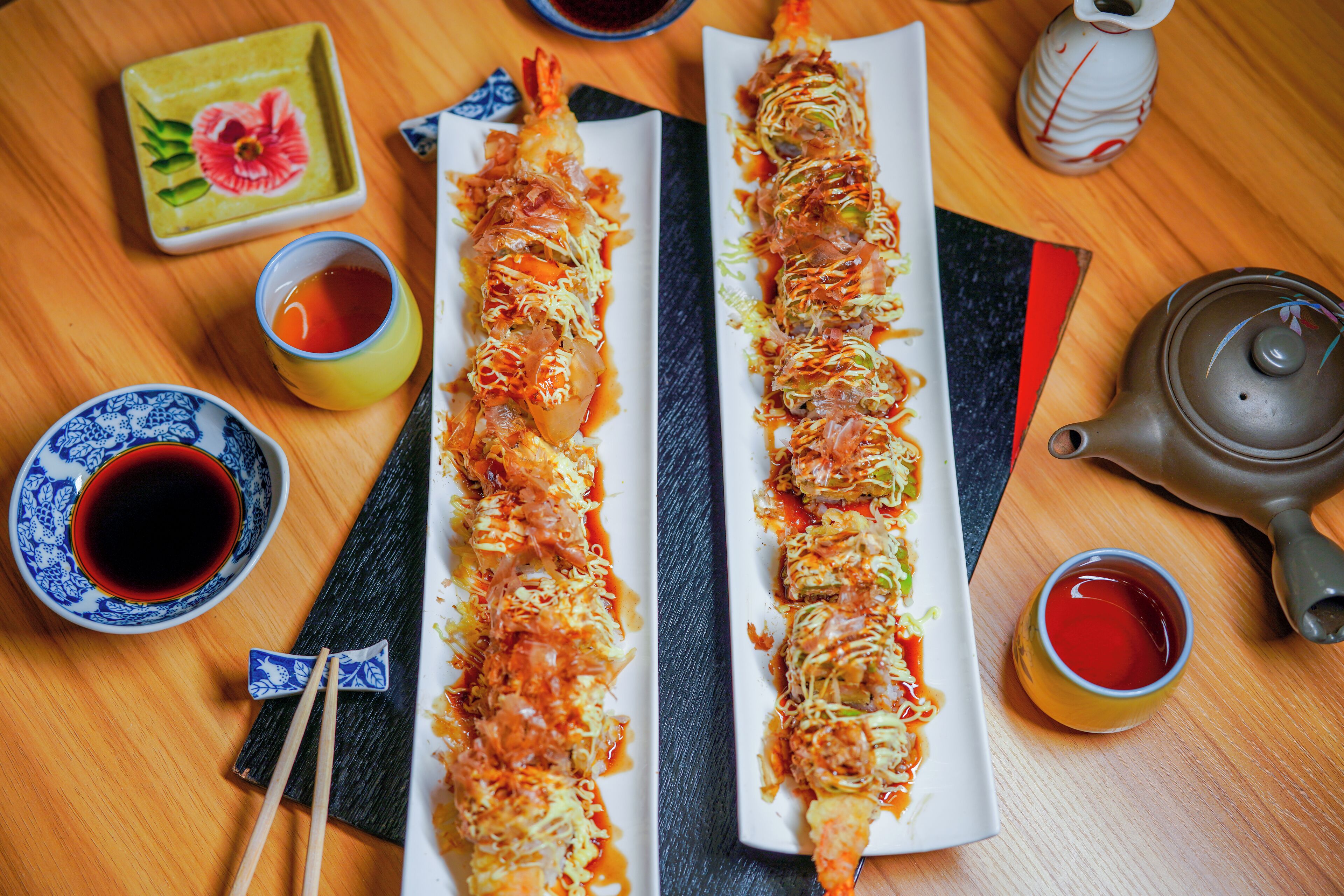 Two long dishes of food on rectangular plates, accompanied by small bowls of sauce and a teapot on a wooden table.