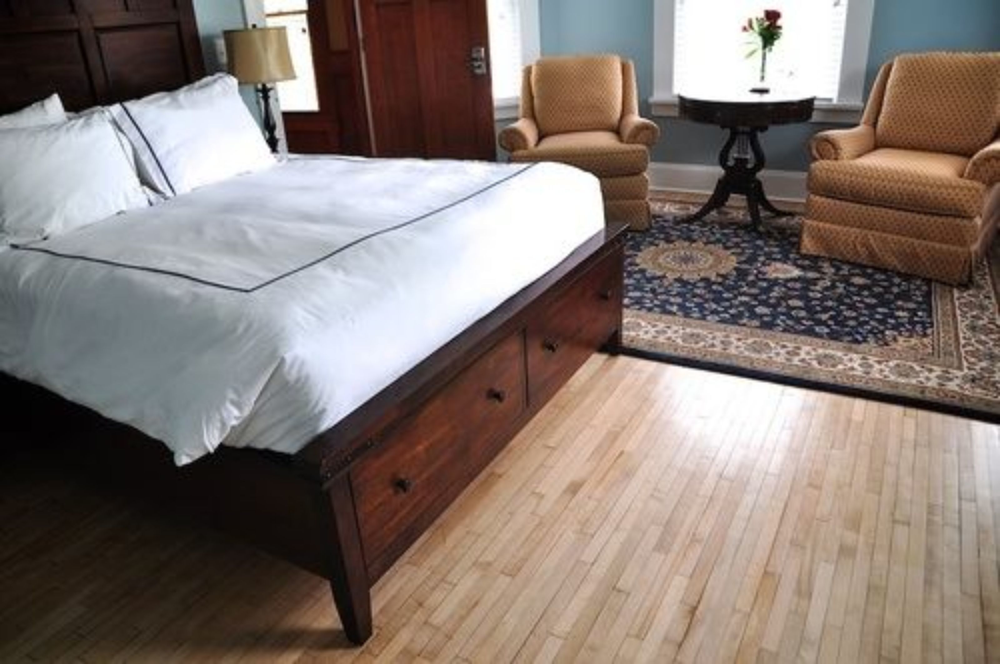 Un dormitorio con una cama grande, cabecera de madera, dos sillones, una mesita redonda y una alfombra estampada.
