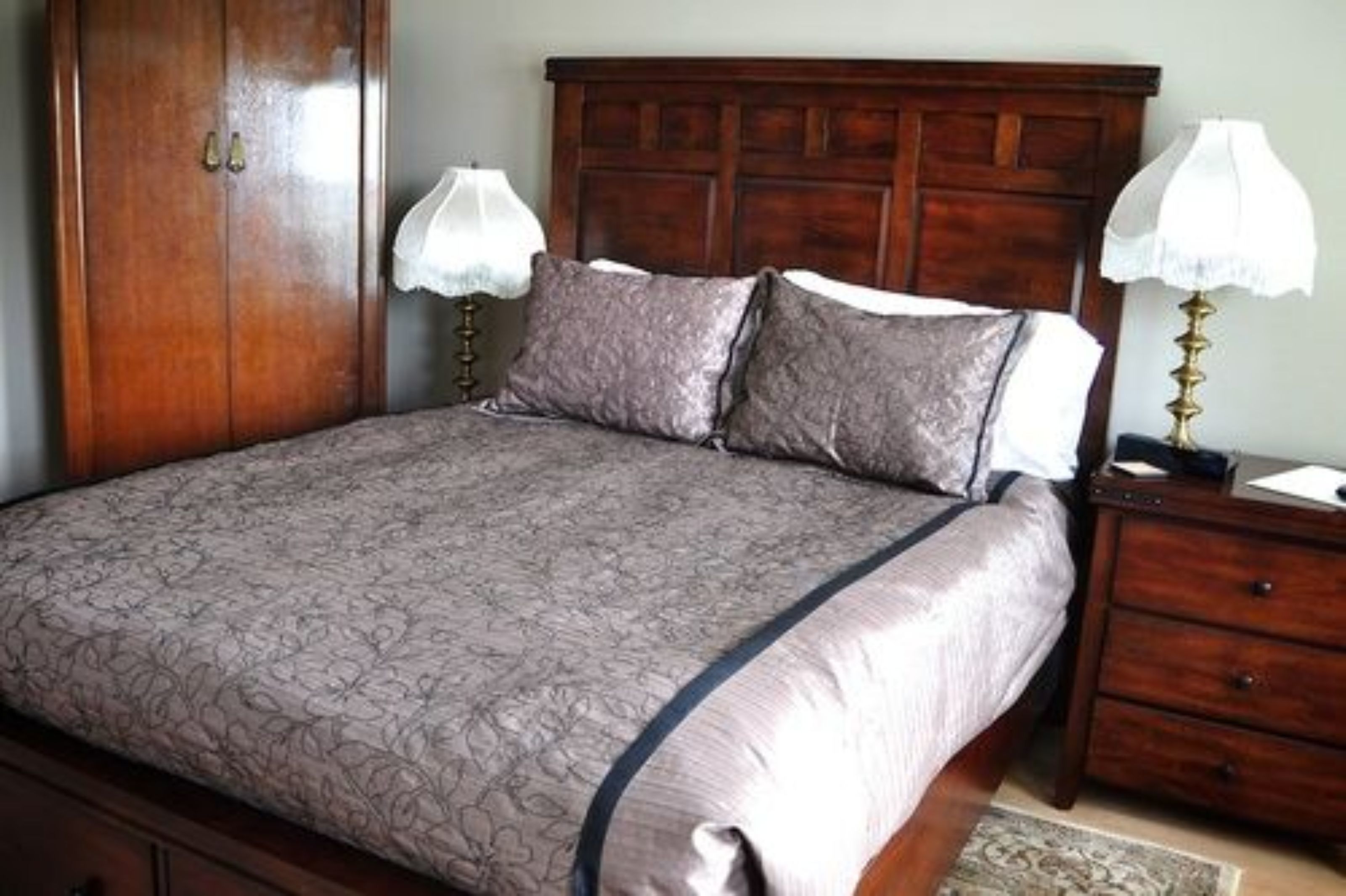 Een slaapkamer met een houten bedframe, een grote garderobe, twee nachtkastjes met lampen en een bed met een decoratief sprei.