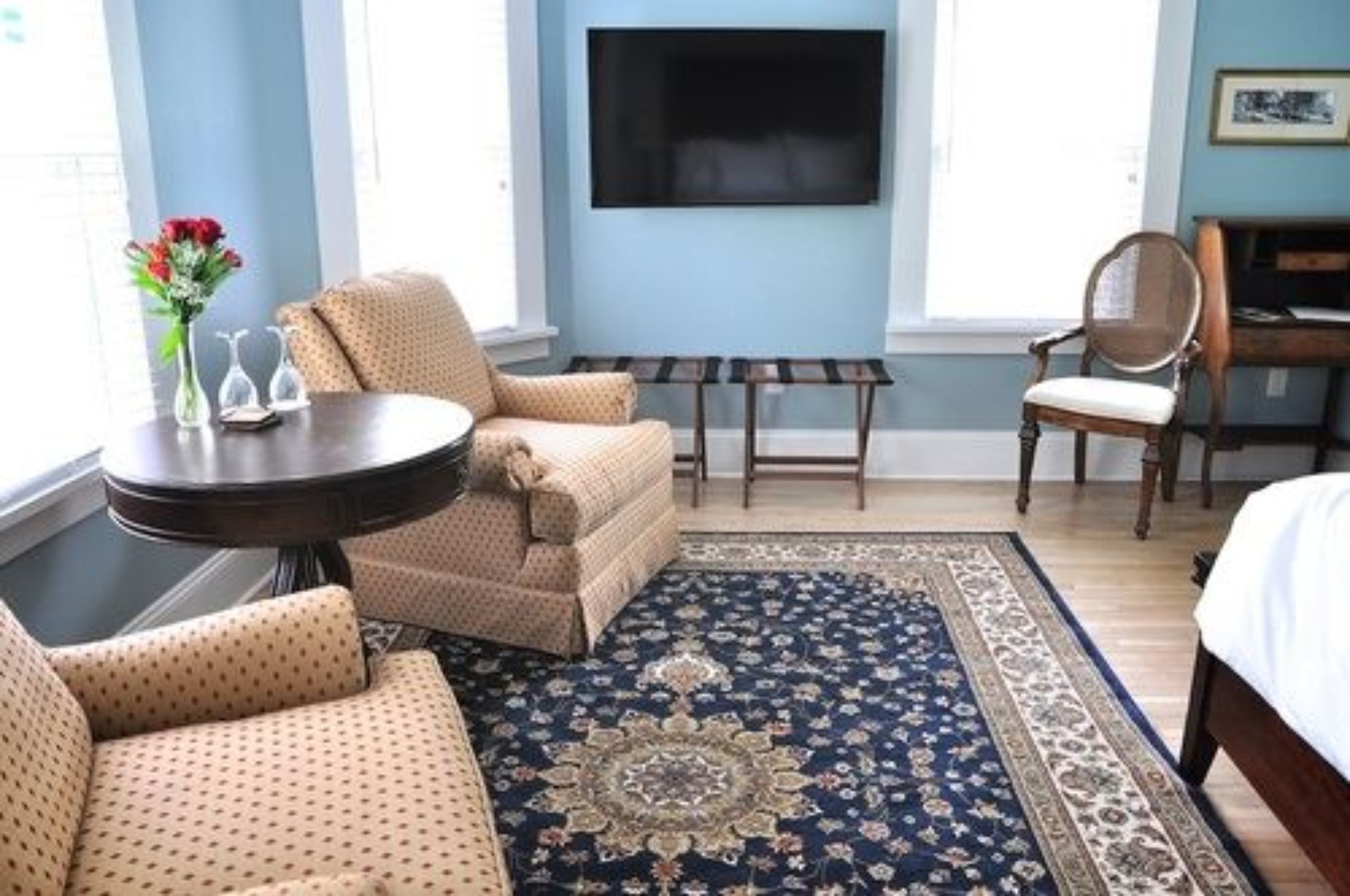 Un cuarto de hotel con una cama grande, un sofá, una mesita con un jarrón de flores, un televisor montado en la pared y una alfombra estampada.