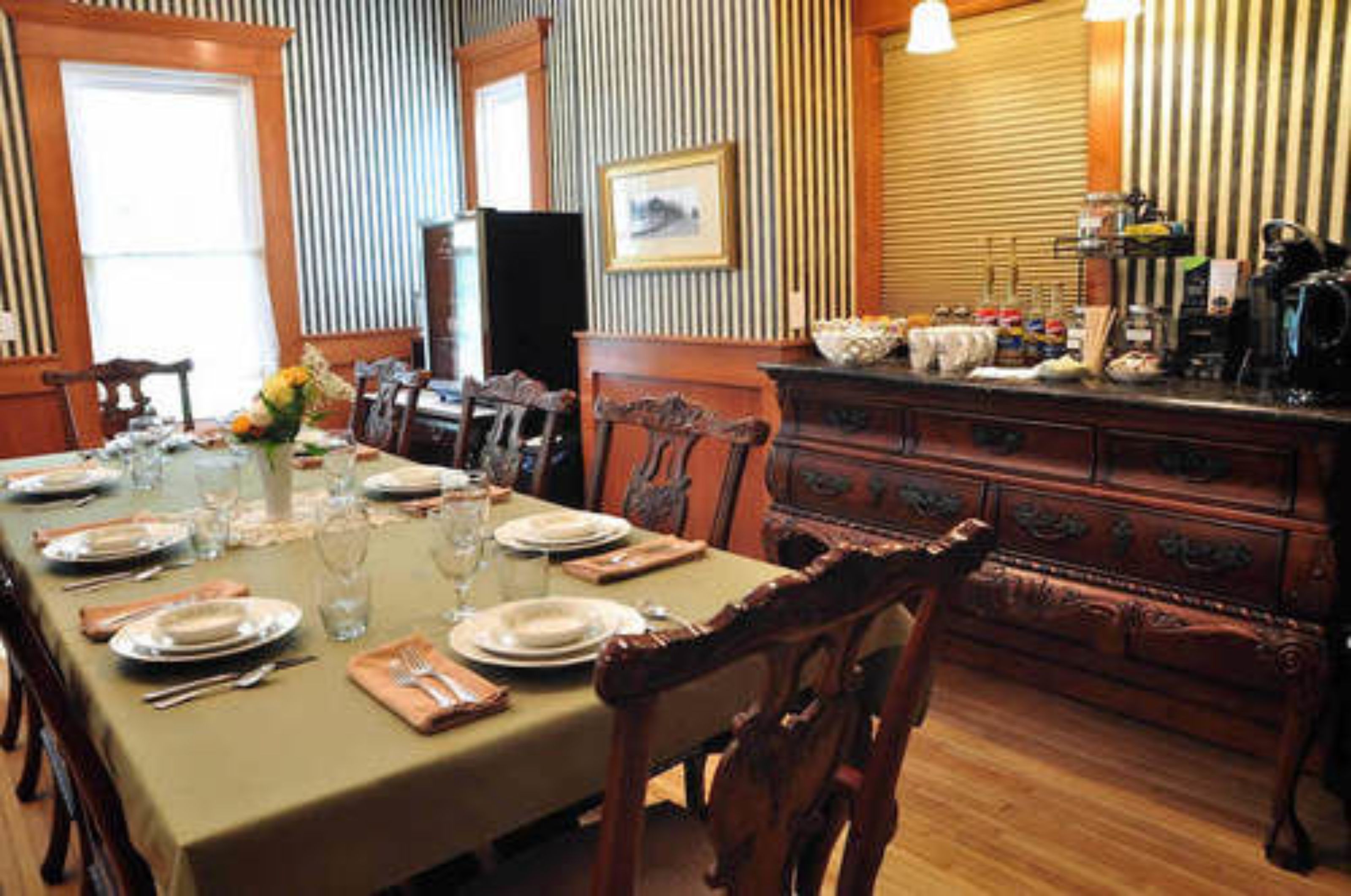 A dining room with a long table set for a meal, wooden chairs, a wooden cabinet with drawers, and a framed picture on the wall.