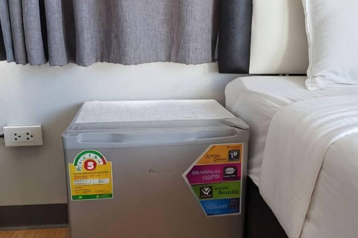 A hotel room with a bed, a white wall, a power outlet, and a trash bin with recycling stickers.