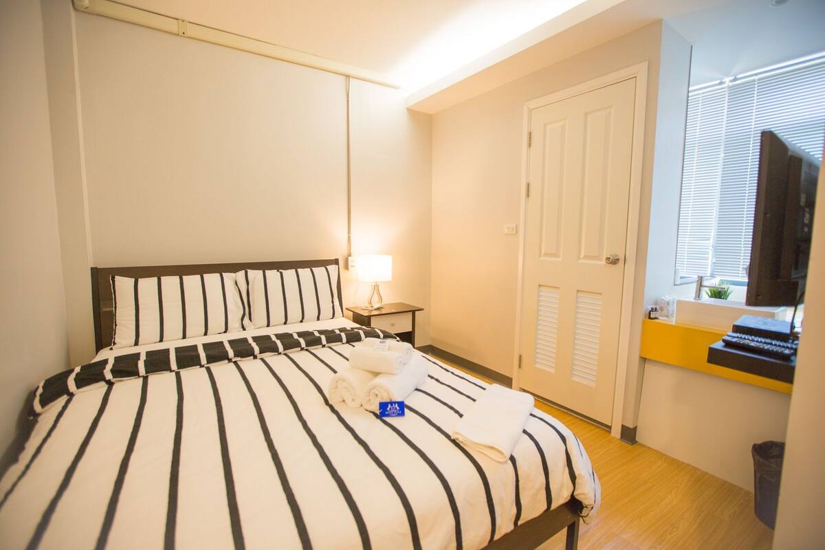 A hotel room with a bed, a bedside table with a lamp, a door, and a window with blinds.
