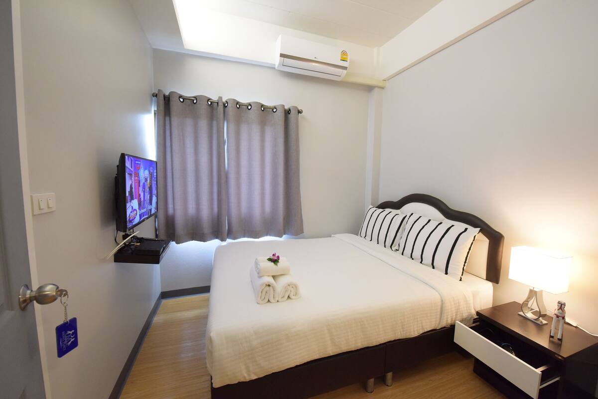 A neatly made bed with white linens and a folded towel, a bedside lamp, a flat-screen TV on a stand, and an air conditioning unit mounted on the wall.