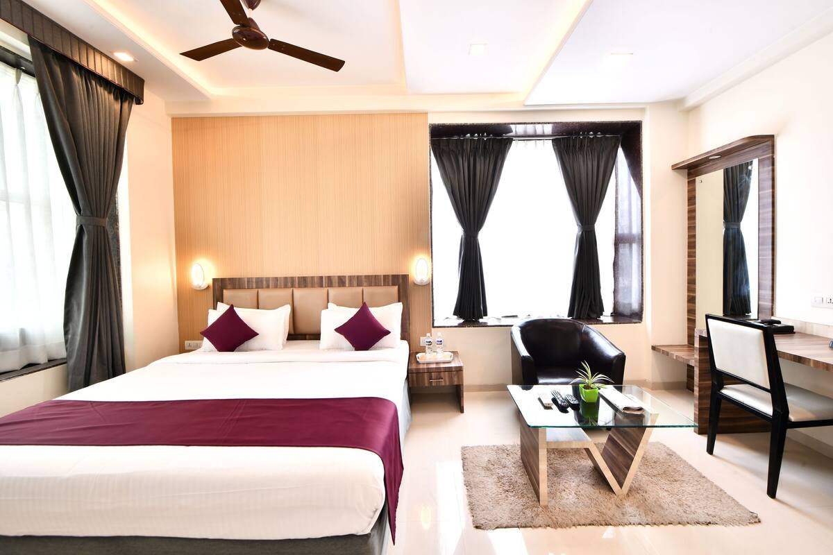 A hotel room with a large bed, a desk, a chair, a ceiling fan, and a large window with curtains.