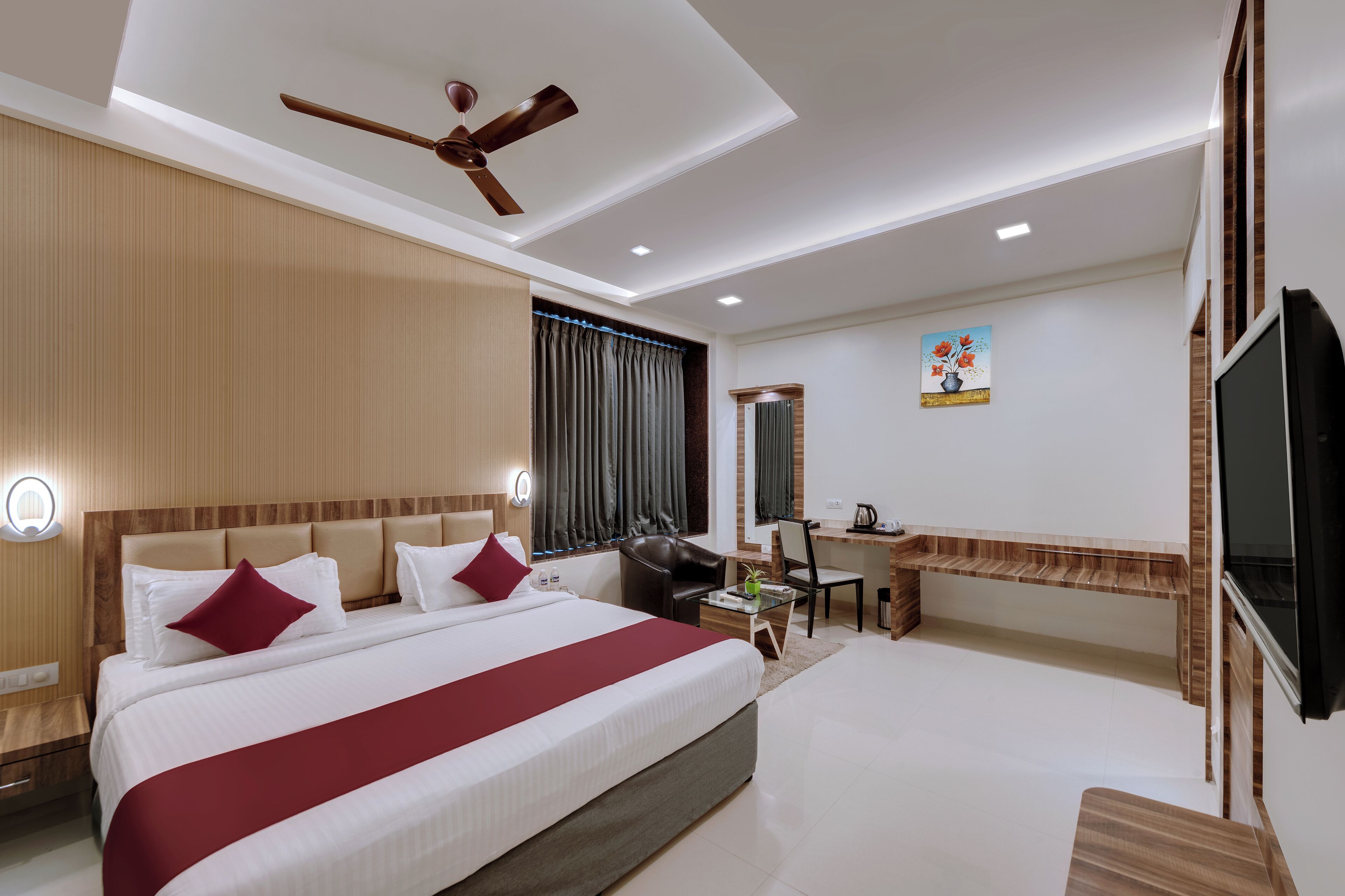 A modern hotel room with a large bed, a desk, a chair, a television, and a ceiling fan.