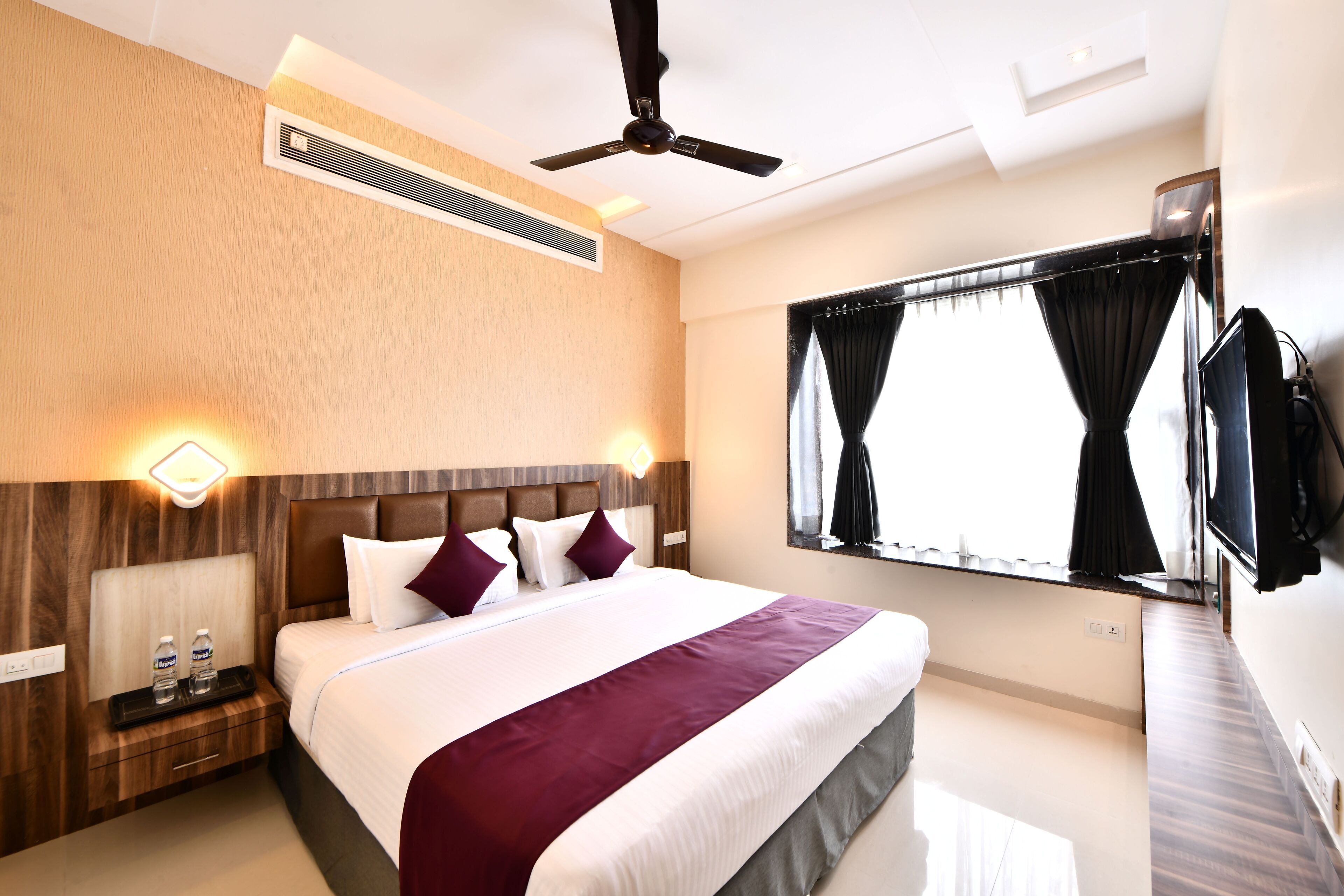 A hotel room with a large bed, a ceiling fan, a TV, and a window with curtains.