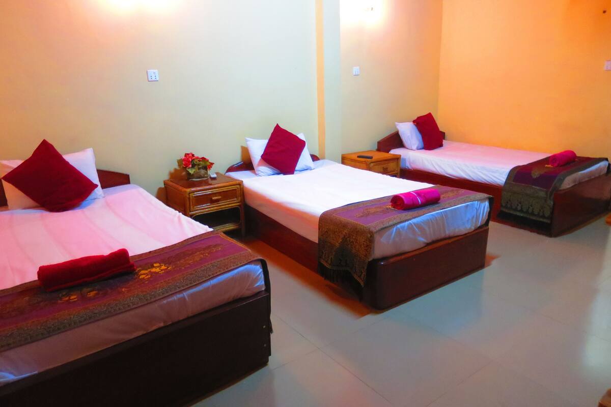 A hotel room with three single beds, each with a red pillow and a decorative headboard. There is a wooden nightstand with a vase of flowers between the beds.