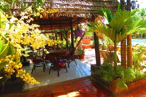 A covered outdoor area with a table and chairs, surrounded by potted plants and a pool. - Kampot Guesthouse (Kampot)