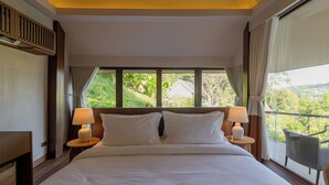 A hotel room with a large bed, two bedside lamps, a view of greenery outside, and a balcony chair.