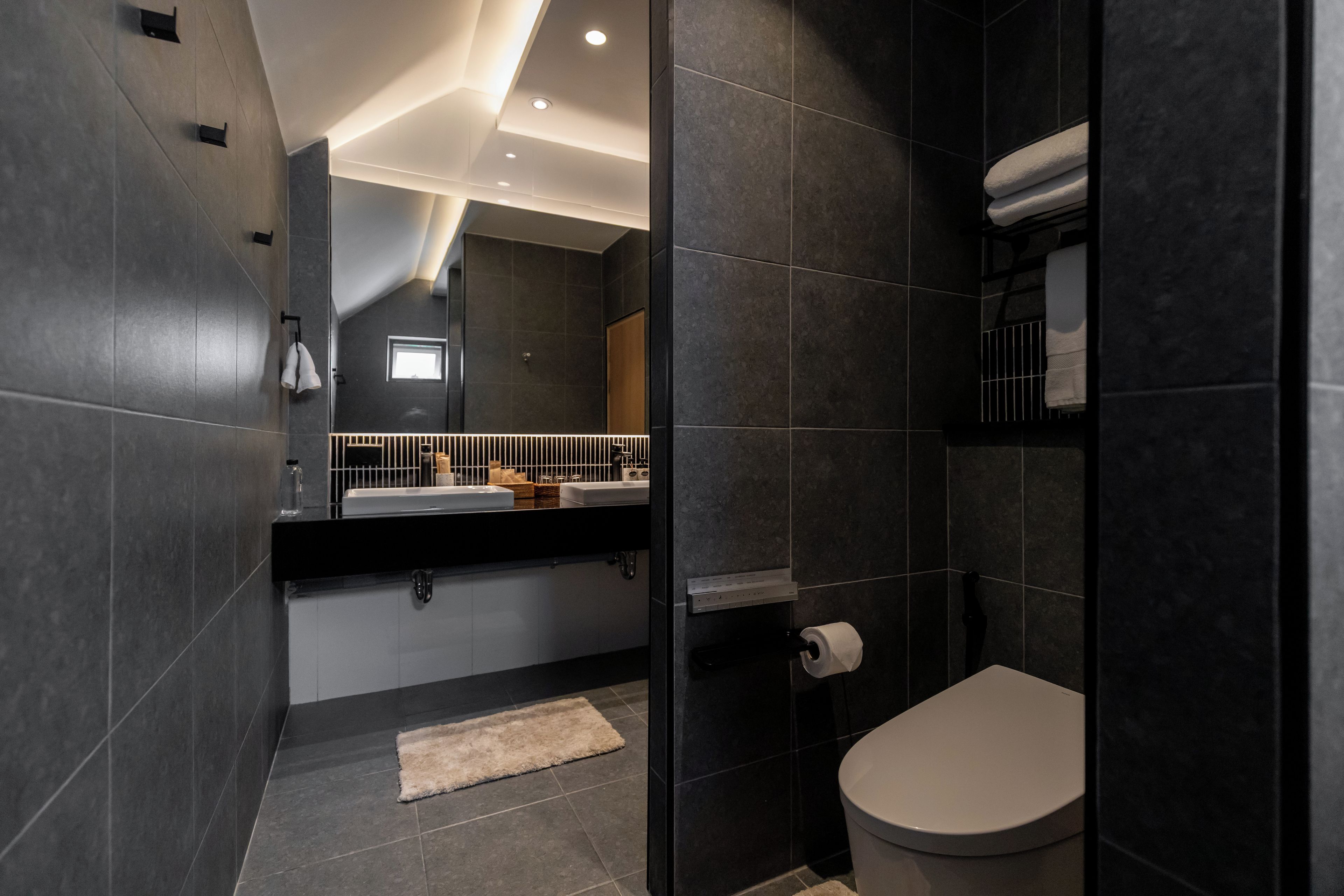 Een moderne badkamer met donkere tegels, een wit toilet, een handdoekhouder en een spiegel.