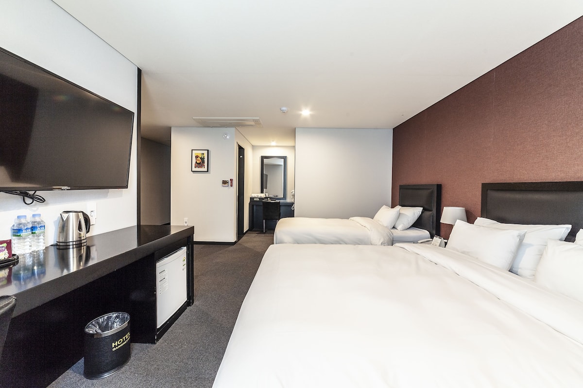 Hotelkamer met flatscreen-tv, bureau met waterkoker, prullenbak, twee bedden en een in de achtergrond zichtbare badkamer.
